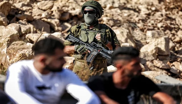 An Israeli soldier stands guard as Palestinians gather in protest at the site of a new road under construction for the use of Israeli settlers near the Palestinian village of Beit Ur